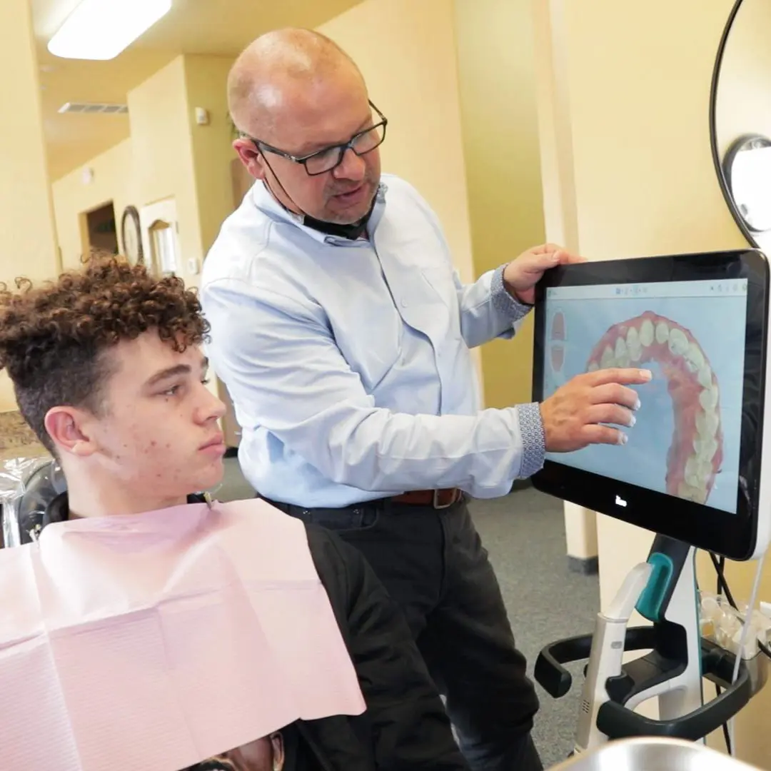 Dr. Gerken explains a digital image of teeth on a screen to a seated patient wearing a pink dental bib. The setting is a well-lit dental office.
