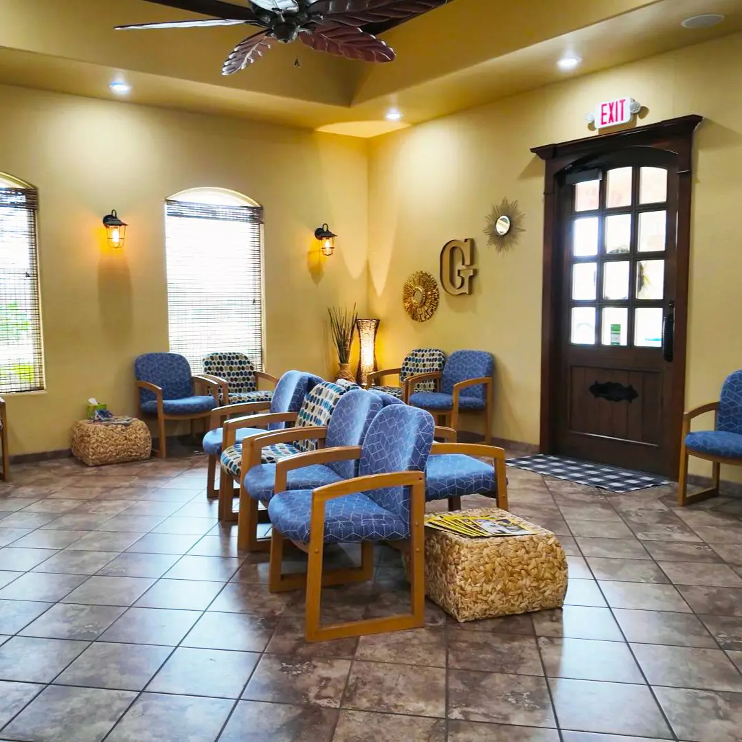 A waiting room with blue cushioned chairs, woven baskets, and tile floor. Sunlight streams through windows, creating a warm, inviting atmosphere.