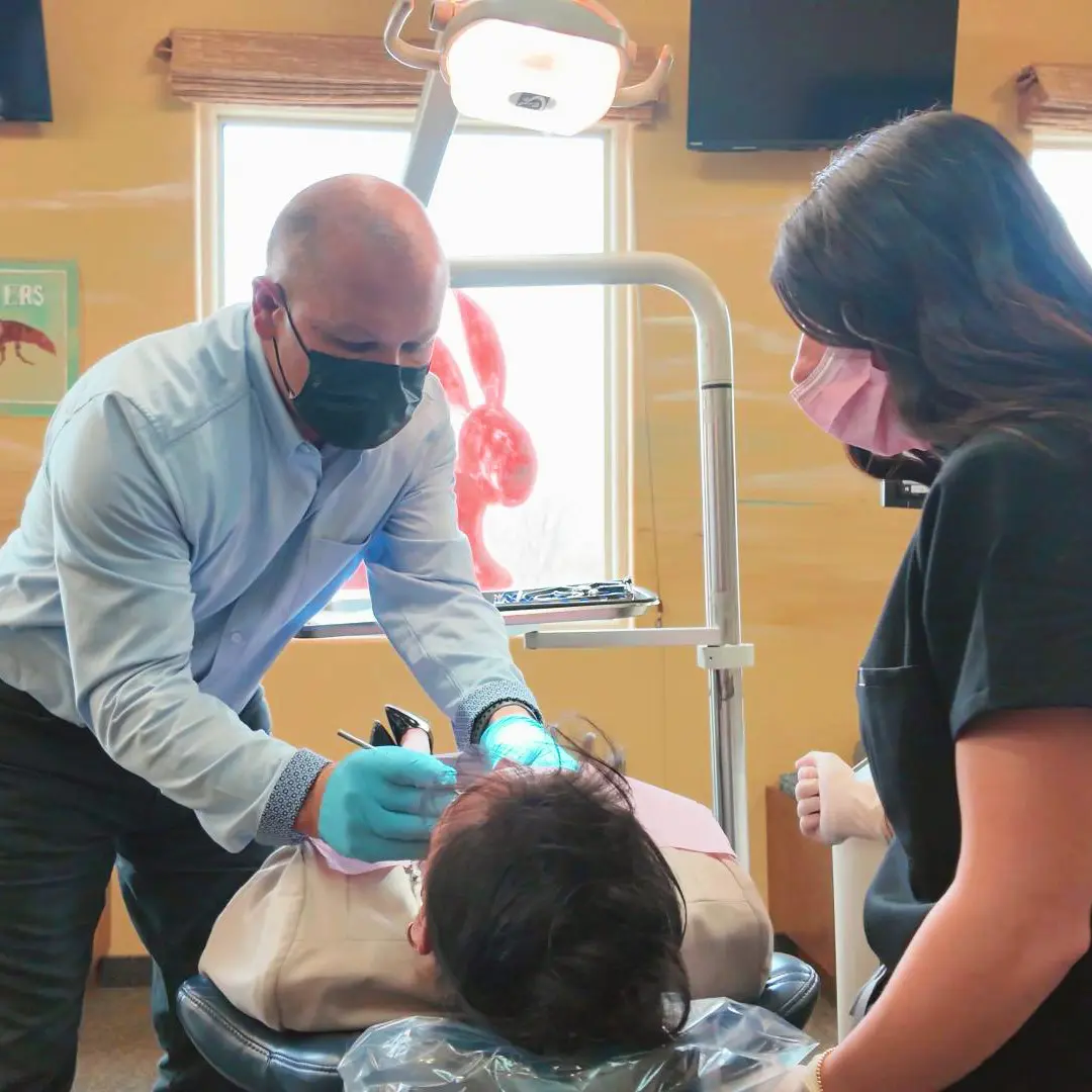Dr. Gerken in a light blue shirt and black mask, examines a patient in a dental chair, assisted by a nurse in black scrubs and a pink mask in a well-lit room.
