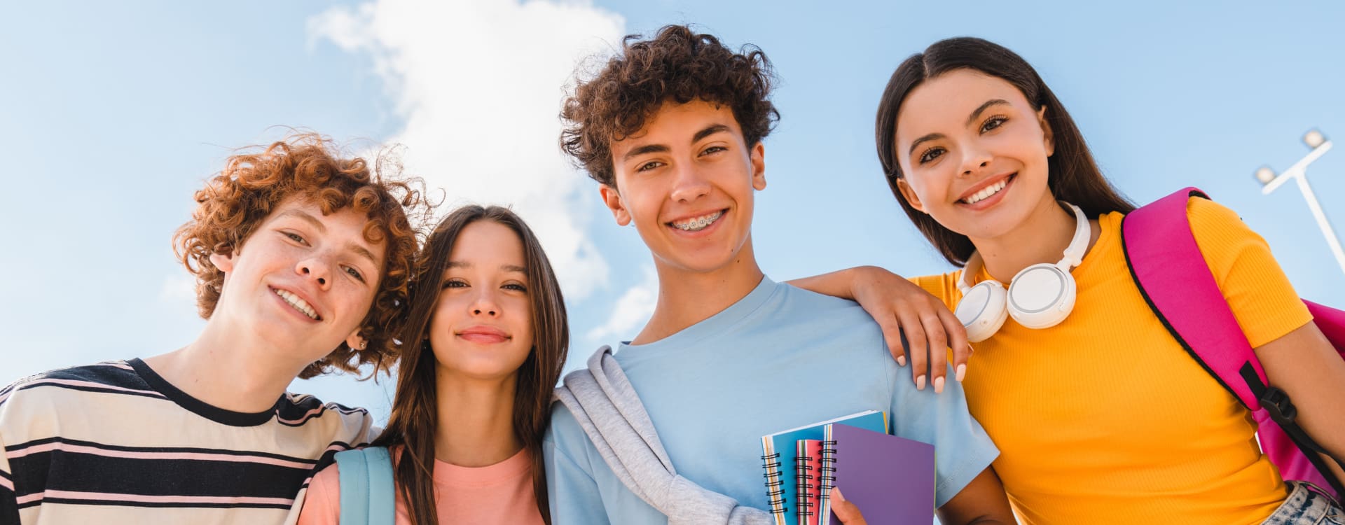 Four smiling teens stand close, arm in arm, under a clear blue sky. They hold notebooks and backpacks, exuding a cheerful and friendly vibe.