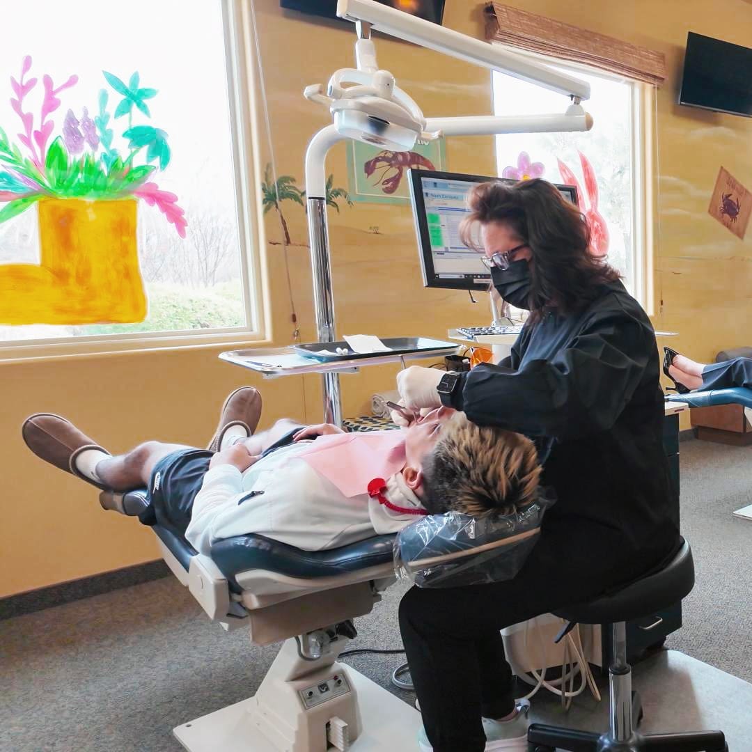 A dental hygienist in a black uniform, wearing a mask, works on a patient reclining in a dental chair. Sunlight filters through a window with colorful painted flowers, creating a calm, cheerful atmosphere.